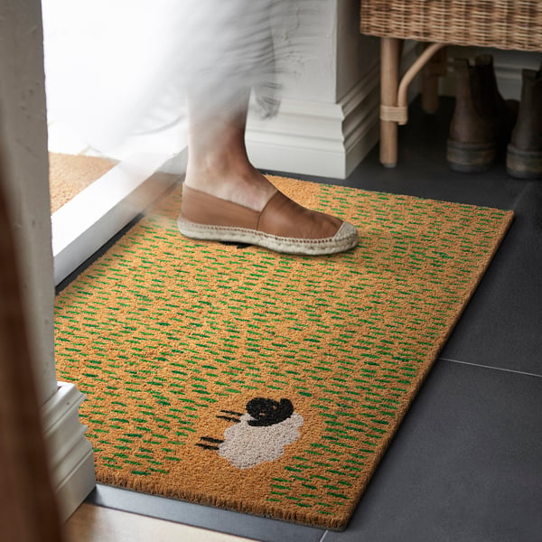 Personen trampar på dekorativ dörrmatta med fårdesign, vilket hindrar jord från att komma in i huset.