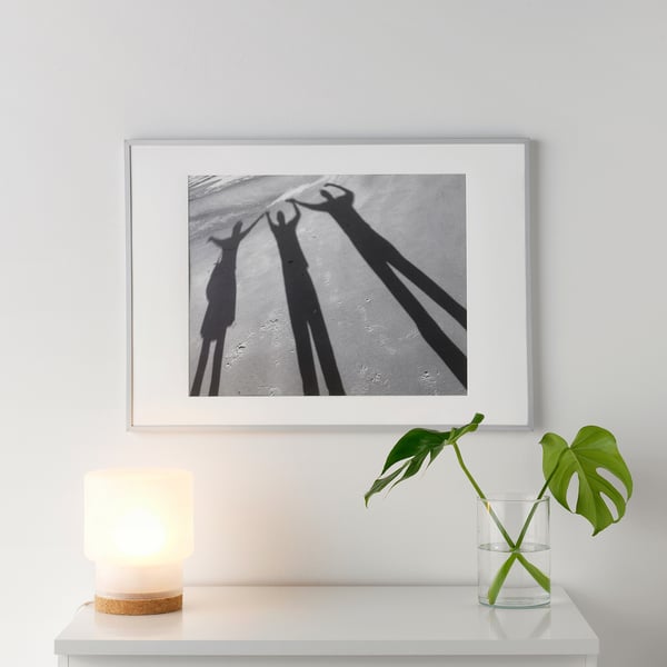 En vit tavelram och svartvita skuggor på en vit hylla. Bredvid den en liten tänd lampa och en glasvas med en grön växt.