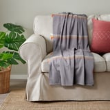 MÄVINN plädfilt, ljusgrå med röda och orange accenter, draperad på en beige soffa, nära en stor krukväxt på vävd matta.