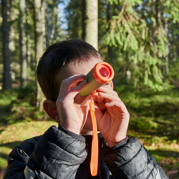 Ett barn håller i ett orange teleskop och utforskar naturen i ett skogsområde. Teleskopet hjälper till att observera avlägsna föremål och uppmuntrar nyfikenhet utomhus.