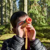 Ett barn håller i ett orange teleskop och utforskar naturen i ett skogsområde. Teleskopet hjälper till att observera avlägsna föremål och uppmuntrar nyfikenhet utomhus.