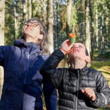 Två personer i en skog, en håller ett barn som använder ett litet grönt och orange teleskop, och tittar upp.