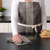 Person i köket, klädd i ett gråtrandigt förkläde, håller en trasa och skär bröd på en svart bänkskiva.