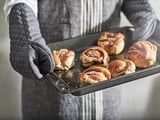 En person i grått förkläde som håller brickan med nybakade kanelbullar.