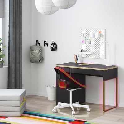 Modern arbetsplats med svart MICKE skrivbord, röd stol, väggförvaring, grå ryggsäck, vit ottoman, färgglad matta.