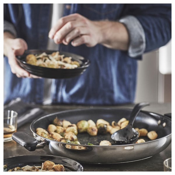 Köksscen: matlagning med svart panna och spatel, stekta grönsaker på spisen, verktyg och vattenglas på bänken.