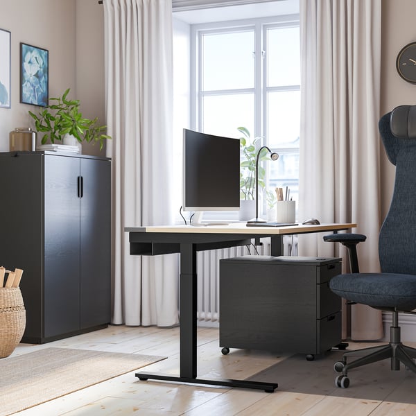 Skrivbordet MITTZON: modern svart design, justerbar höjd, rymlig arbetsplats nära stora fönster, grå stol, förvaring och växter.