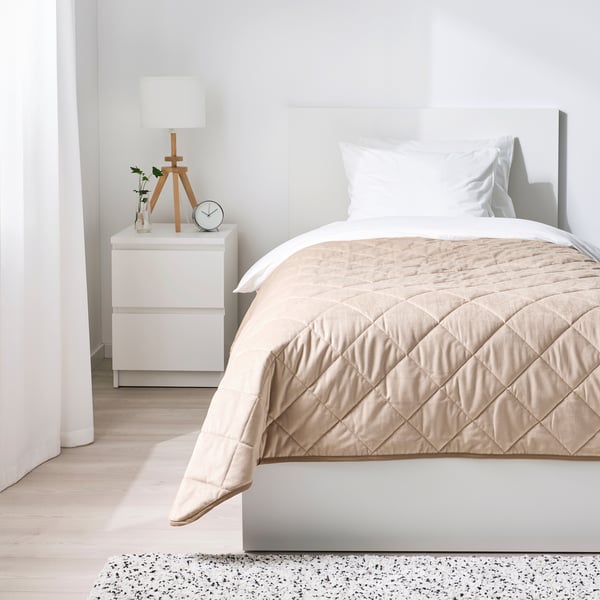 Minimalistiskt sovrum med vita väggar, beige överkast, nattduksbord, lampa och klocka. Mysig, lyxig känsla med mjukt, quiltat överkast.