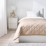 Minimalistiskt sovrum med vita väggar, beige överkast, nattduksbord, lampa och klocka. Mysig, lyxig känsla med mjukt, quiltat överkast.