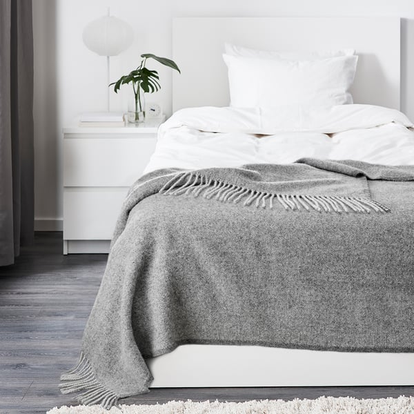 Minimalistiskt sovrum med en vit säng med en mysig, strukturerad grå ullfilt som är snyggt vikt vid fotändan.