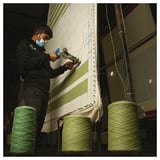 Man tuftande grön och beige matta med verktyg, omgiven av garnspolar i industriell verkstad.