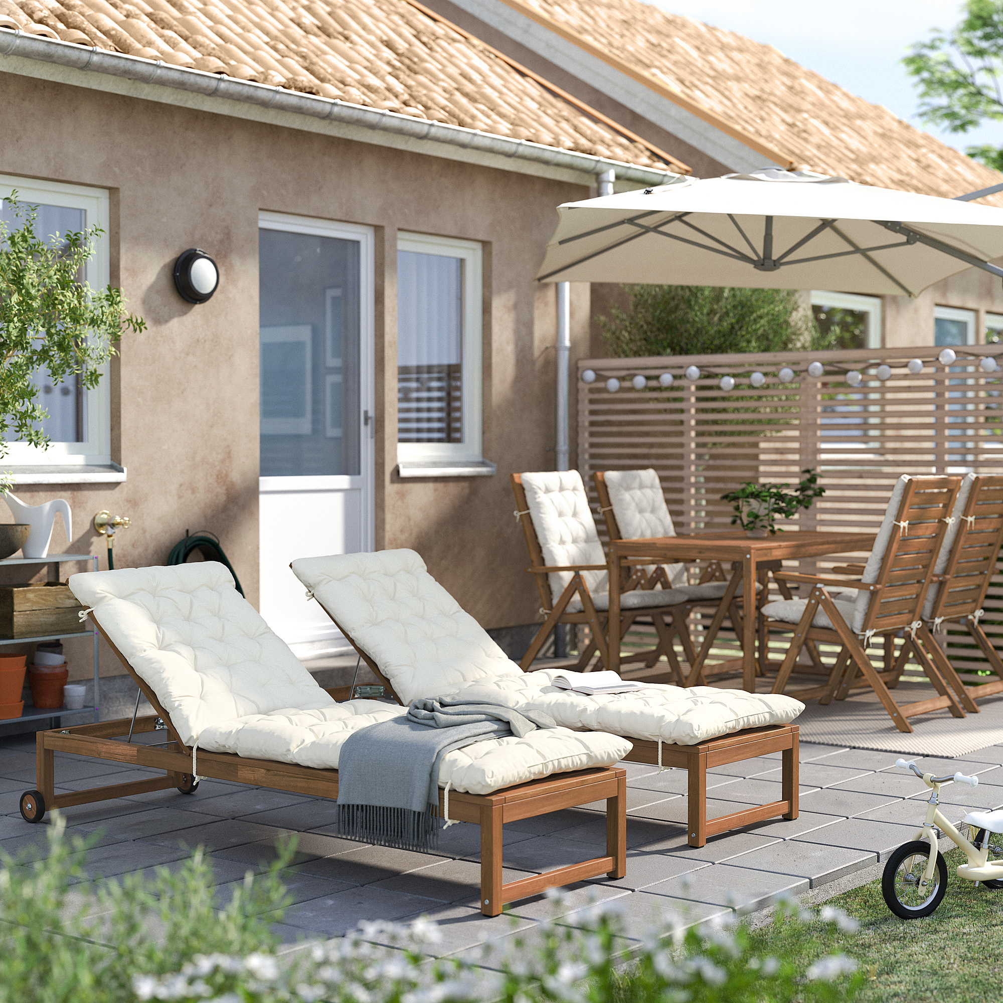 Uteplats på bakgården med NÄMMARÖ vita vadderade vilstolar, träbord, stolar och stort parasoll nära huset.