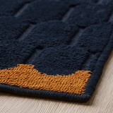 Mörkblå texturerad matthörna med tätt, senapsfärgat garnmönster och robust baksida på trägolv.