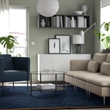 Modernt vardagsrum med NÖVLING-blå matta, grå soffa, blå stol, soffbord i glas, vitt skåp, böcker, vaser och växter.
