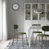 Modern matplats med gröna stolar, vitt bord, silverljus, randig matta, klocka, bokhylla.