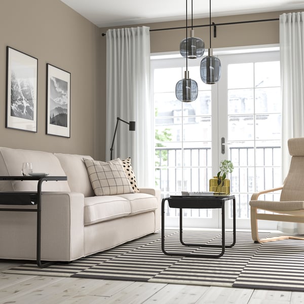 Modernt vardagsrum med ljusbeige soffa, randig matta, tre svarta taklampor, svart sidobord och en balkong.