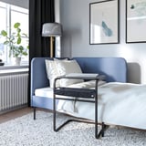 Blå soffa med vita kuddar, bärbar dator på flyttbart bord, grå matta, växt, lampa.