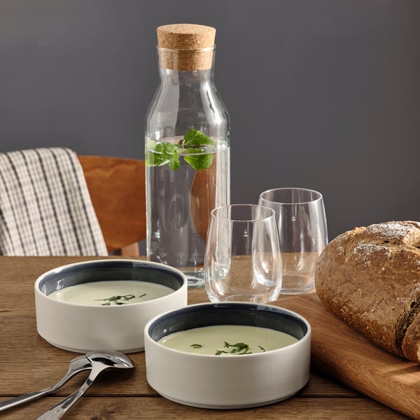 Två keramiska skålar med soppa, två glas, en glasburk med vatten med örter, trästol, randig servett, brödlimpa på ett träbord.