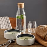 Två keramiska skålar med soppa, två glas, en glasburk med vatten med örter, trästol, randig servett, brödlimpa på ett träbord.