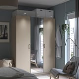 Modernt sovrum med en beige PAX-garderob, spegeldörrar och grå väggar. En hängande växt, en stol med en filt och en säng delvis synlig.