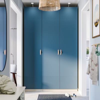 En stor blå PAX garderob med modern design. Dess storlek överstiger närliggande möbler och den har en elegant, räfflad struktur och guldhandtag.