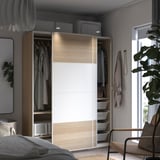 Ett modernt sovrum med en ljus och vit PAX-garderob i trä. Den har skjutdörrar, en öppen sektion med kläder och nätkorgar för förvaring.