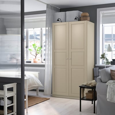 Ljust sovrum med stor beige PAX-garderob, vita möbler, grå soffa och krukväxter, med stora fönster.