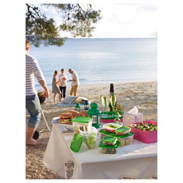 Strandpicknick med gröna och rosa matbehållare på en duk. Människor som står nära havet.
