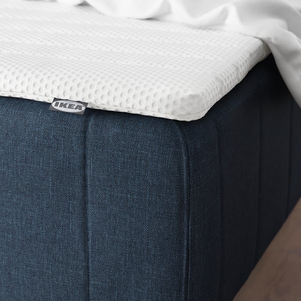 En vit texturerad madrass märkt IKEA på en mörk säng, designad för komfort och stöd.