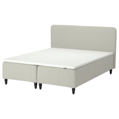 En beige säng RÅHOLMAR med stoppad sänggavel, vit madrass och svarta ben som ger stöd och tryckavlastning.