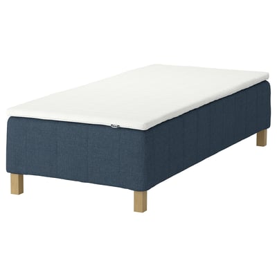 Bilden visar en blå säng RÅHOLMAR med en vit madrass. Stoppat tyg och träben, strömlinjeformad design.