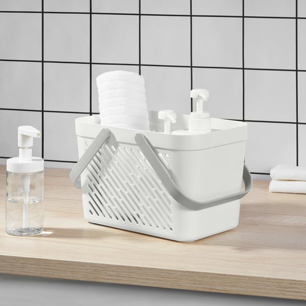 Duschkorg i vit plast med vikbara handtag, staplade med rullade vita handdukar, placerade på bänkskivan i ljust trä bredvid klar tvålpump.