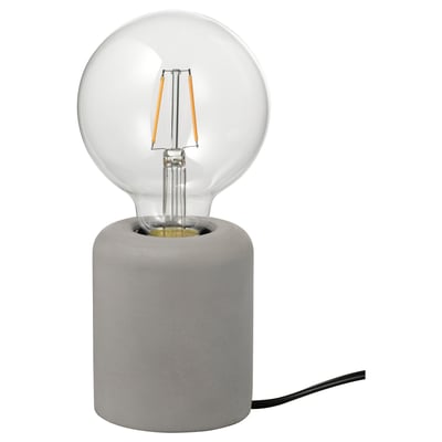 En RÅSEGEL-lampa med en betongfot och en glödlampa i glas som avger varmt ljus.