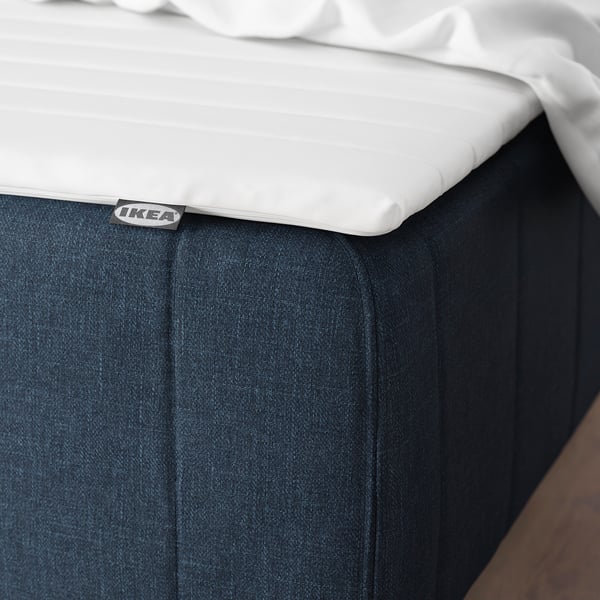 Vit madrass på mörk sängkjol med IKEA-etikett synlig.