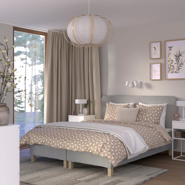 Modern grå säng RENFJÄLLET med geometriska sängkläder, taklampa, nattduksbord, blomsterkonst och stora fönster.