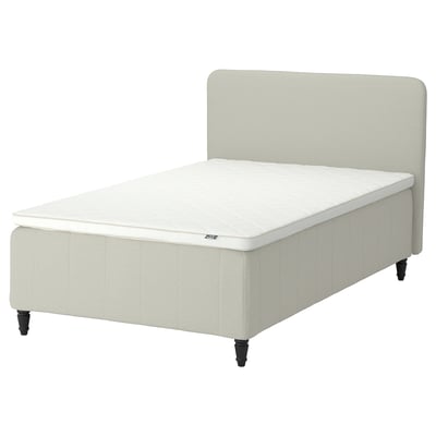 Rishojden-sängen är en beige stoppad ram med vadderad sänggavel och vit madrass. Den har en rektangulär form med avsmalnande träben.