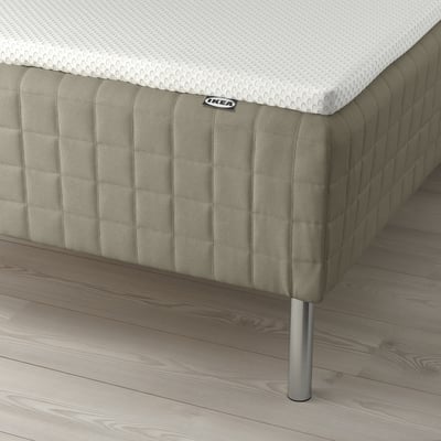 Säng med IKEA SKÅRER madrass på trägolv, stödd av metallben, vadderad i grått tyg.