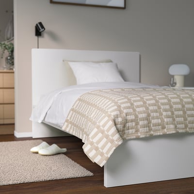 Mysigt sovrum med beige överkast på stor vit säng, neutral inredning, vita kuddar, tofflor på matta vid trägolv.