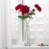 En klar vas med röda konstgjorda blommor på en fönsterbräda. Blommorna har gröna stjälkar och blad.