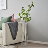 Vas som heter SMYCKA med grön växt nära beige soffa. Högt, klart glas som innehåller naturtrogen konstgjord grönska. Dekorativ, modern heminredning.