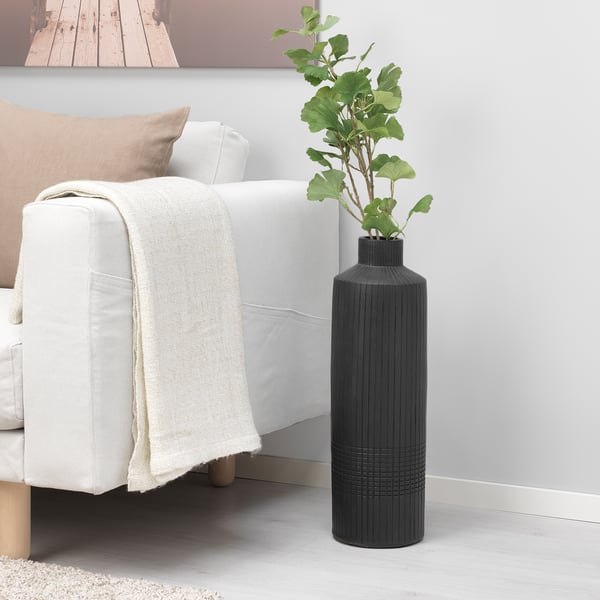 Ett minimalistiskt vardagsrum med en vit soffa, en beige prydnadskudde och filt och en stor svart dekorativ vas med gröna konstgjorda blommor.