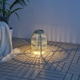 Grön SOLVINDEN golvlampa med rottingmönster på däck, upplyst med växt och tyg.