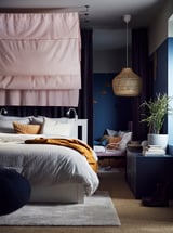 Städat sovrum med SONGESAND vit säng, rosa gardiner, mörkblå väggar, hängande korglampa och växter.