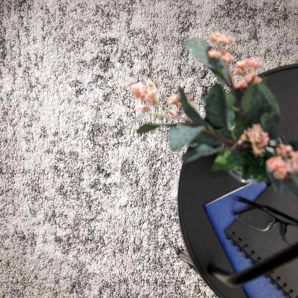 Ovanifrån av en grå, rund matta med en vas som rymmer rosa blommor och gröna blad på ett bord.