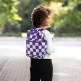 En person bär en lila och vitrutig ryggsäck med en sidoficka för vattenflaska, lämplig för skolan med åldern 7+.