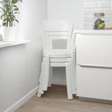 Två vita plaststolar staplade i modernt kök. Lätt, elegant, med skålsits och rundat ryggstöd. Bredvid disk med växt- och fruktskål.