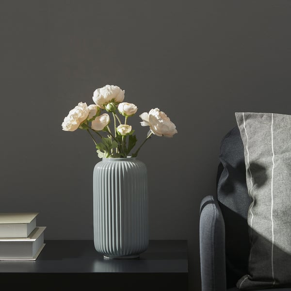 En modern vas med vita blommor står på ett bord bredvid en trave böcker och en stol med en filt draperad över den.