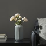 En modern vas med vita blommor står på ett bord bredvid en trave böcker och en stol med en filt draperad över den.
