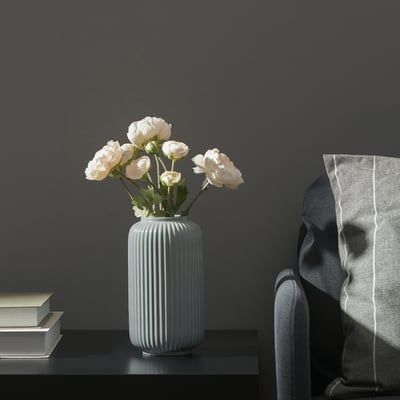 En modern vas med vita blommor står på ett bord bredvid en trave böcker och en stol med en filt draperad över den.
