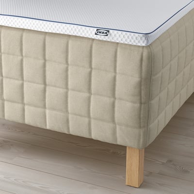 Visar en säng med en ljus träbotten och en texturerad madrass. Synliga ben och en märkesbricka.